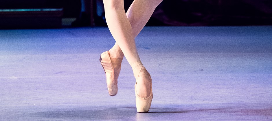 En ballerina som dansar på en scen. Man ser hennes underkropp. Hon är klädd i tajts och tåspetsskor. 