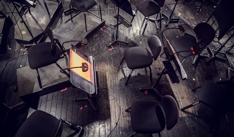 En liten lampa på ett notställ står i mitten på scenen omringat av orkesterns svarta tomma stolar. 